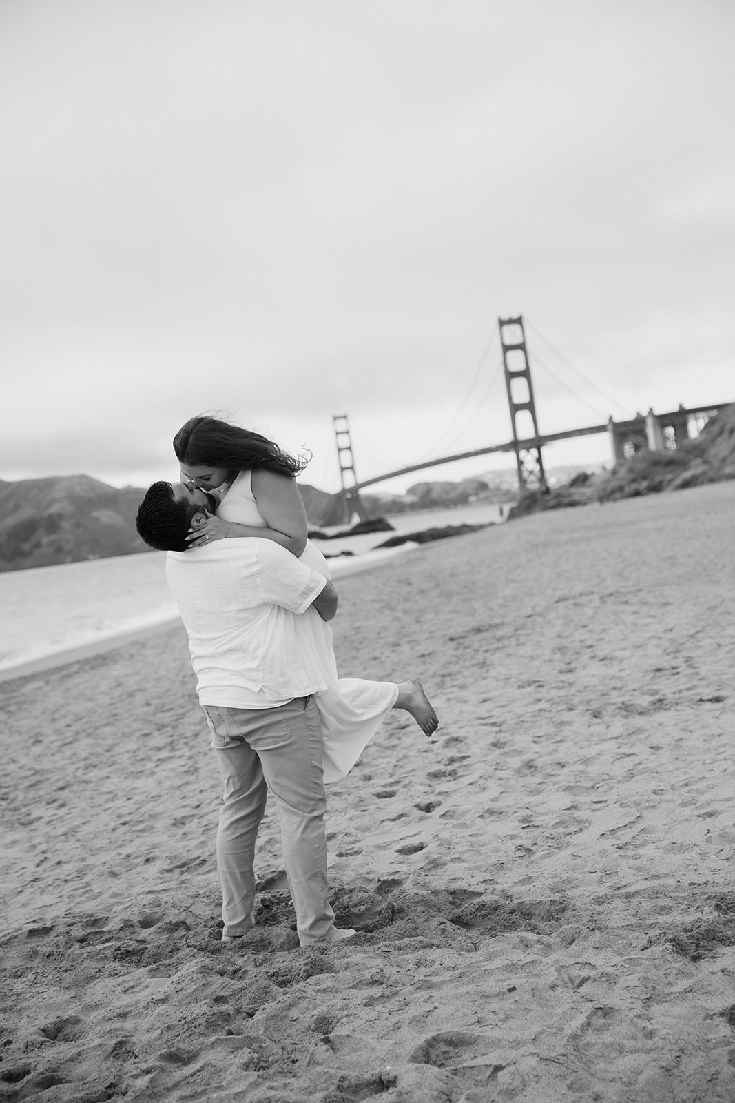 a couple having an engagement session at baker beach in san francisco the man is picking up his partner and they are kissing