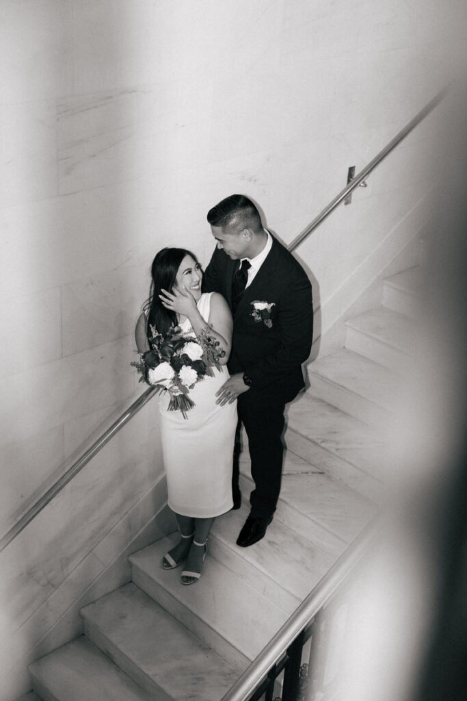 A newlywed couple standing on a staircase together smiling at each other 