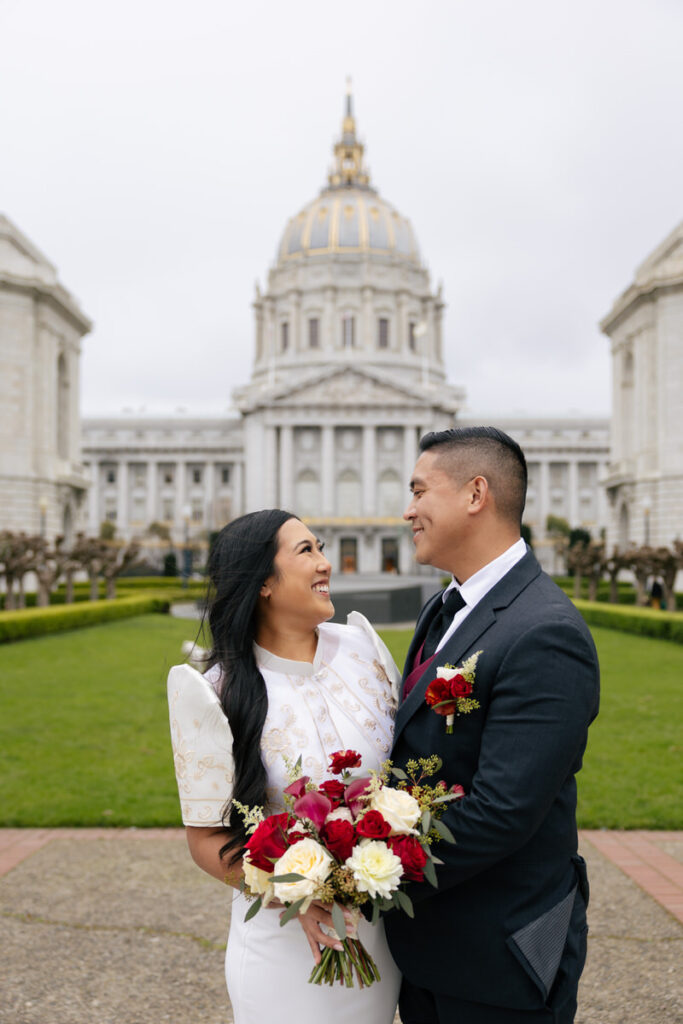 A wedding couple standing in front of the San Francisco City Hall building while one holds a bouquet of flowers 