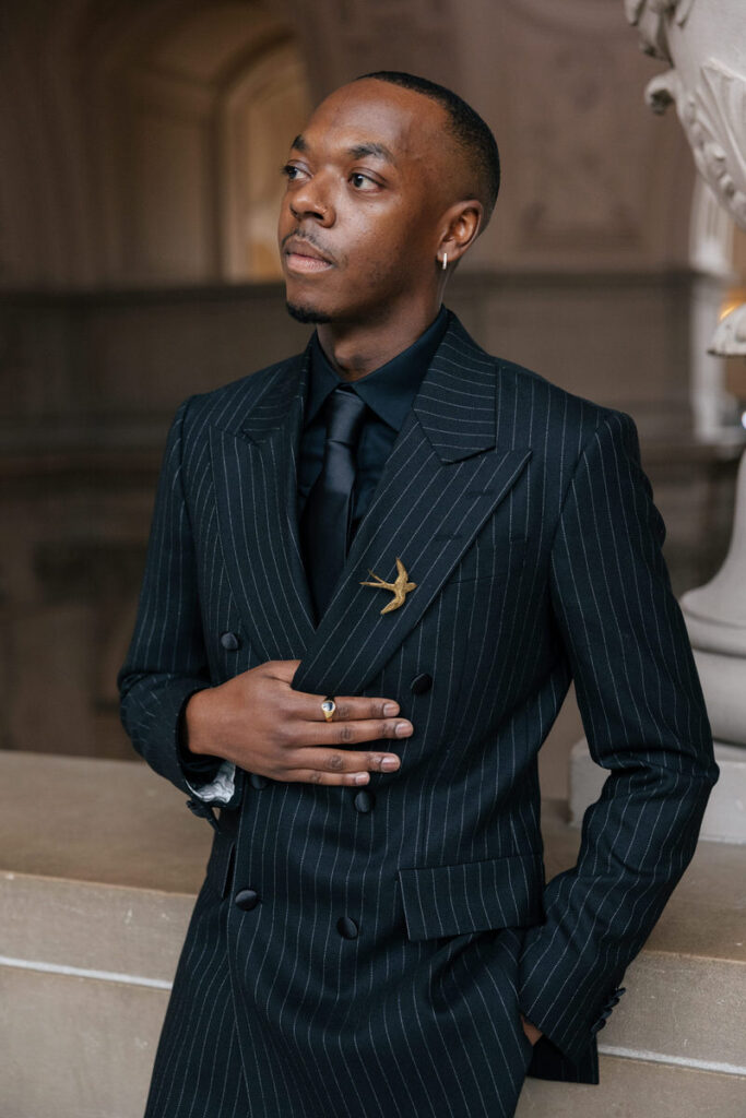 A person in a black suit with one hand on their jacket 