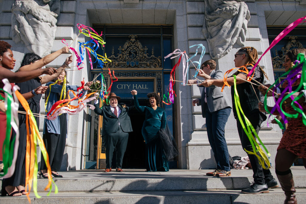 A newlywed couple walking out of City Hall with their wedding guests waving colorful streamers 