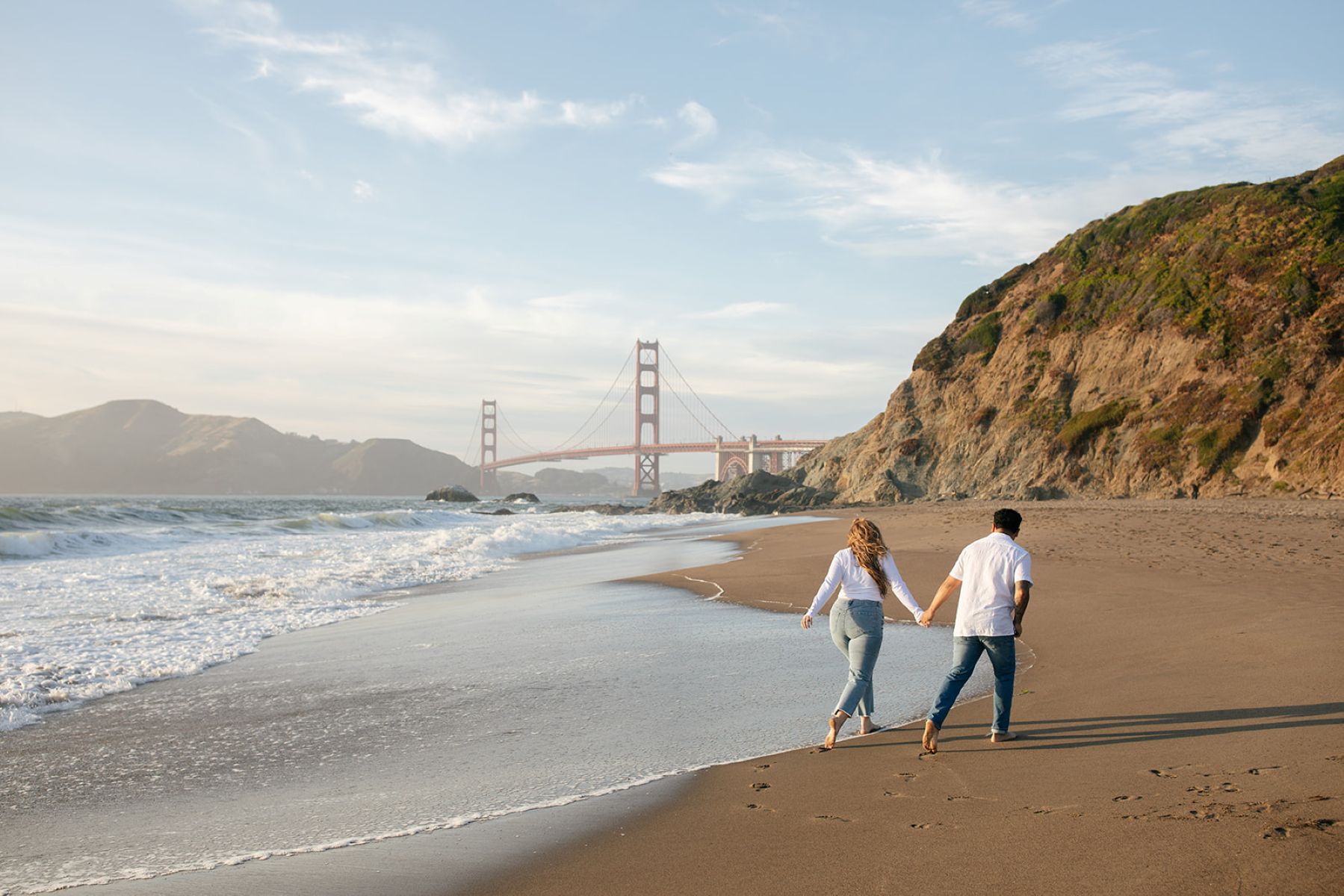 a couple holding hands on the beach taking their san francisco engagement photos at baker beach at golden hour
