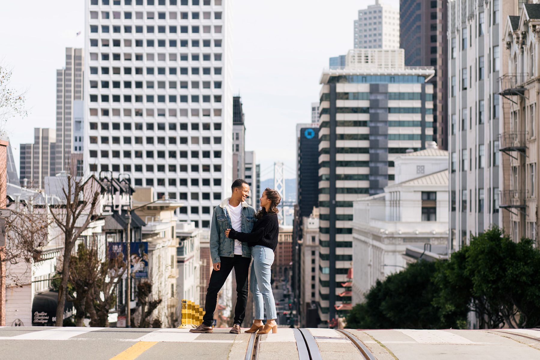 a copule standing on the powell street intersection in san francisco taking engagement photos the woman is hugging her partner and they are both smiling at each other
