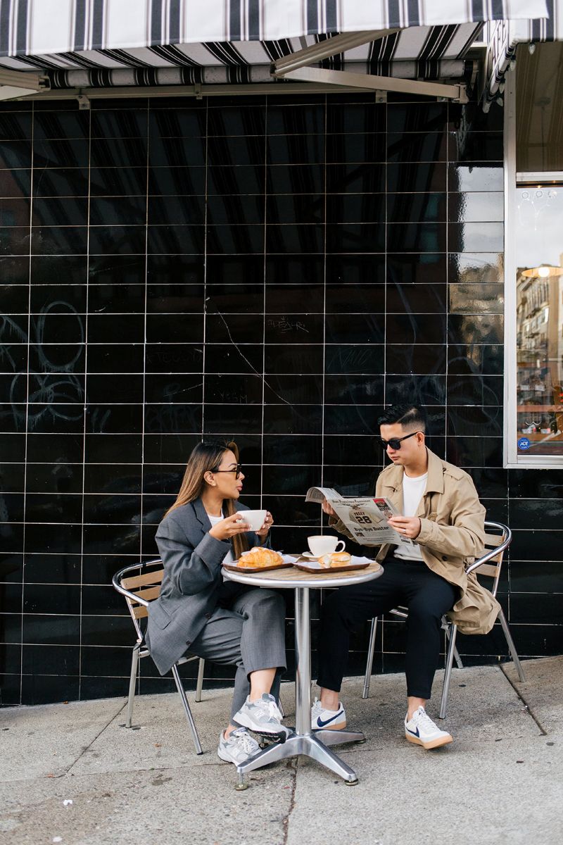 a couple having an engagement session at a coffee shop they are both sitting in chairs outside and the woman is holding a cup of coffee in her hand the man is reading a newspaper