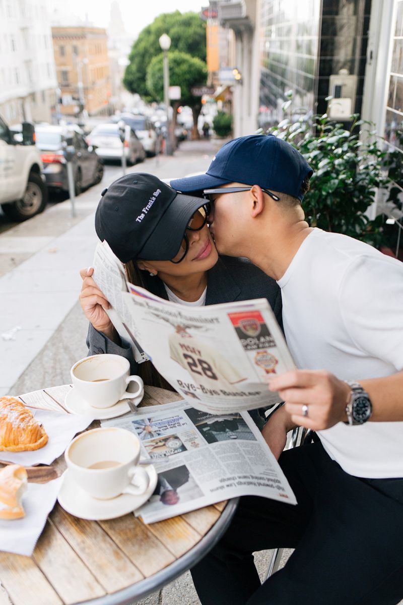 a couple sharing a newspaper and sitting at a table the man is kissing his partner on the cheek they are both wearing hats and sunglasses and taking engagement photos in san francisco