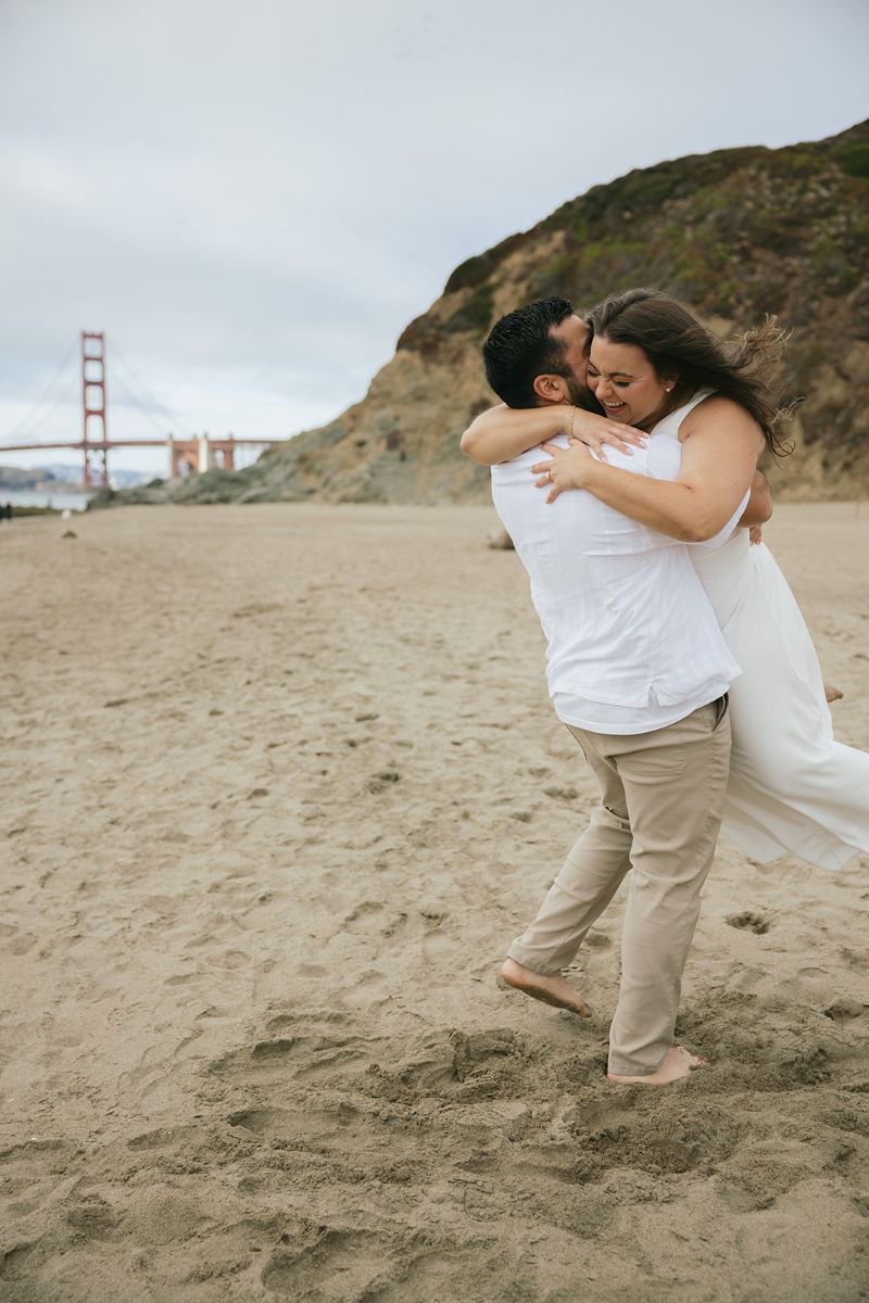 a couple having an engagement session at baker beach in san francisco the man is picking up his patner and kissing the side of her cheek wile she is laughing