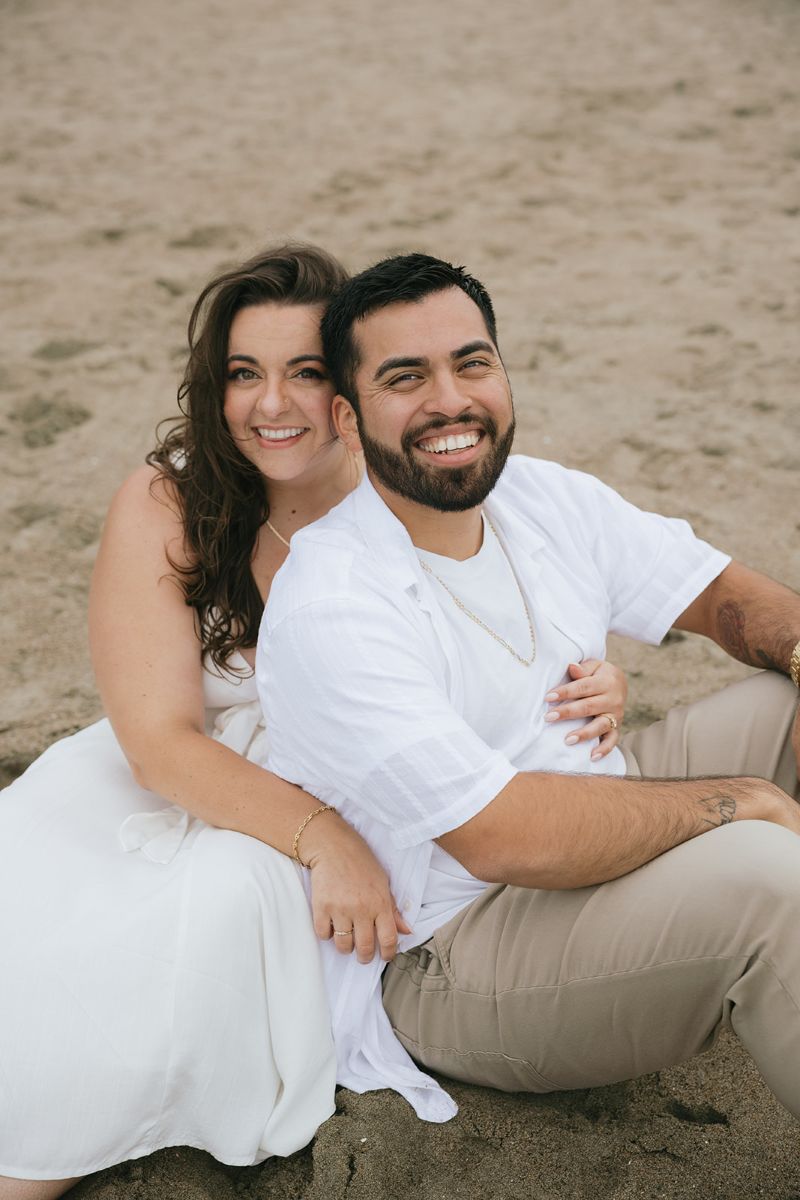 a couple sitting on a beach in the sand taking engagement photos they are both smiling and the woman has her arm wrapped around her partner