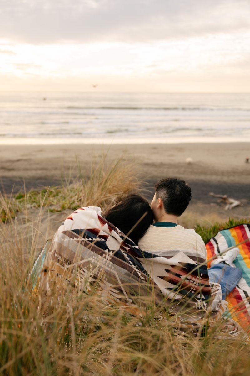 a couple wrapped in a blanket sitting next to each other at the beach one has their head on their partner's shoulder