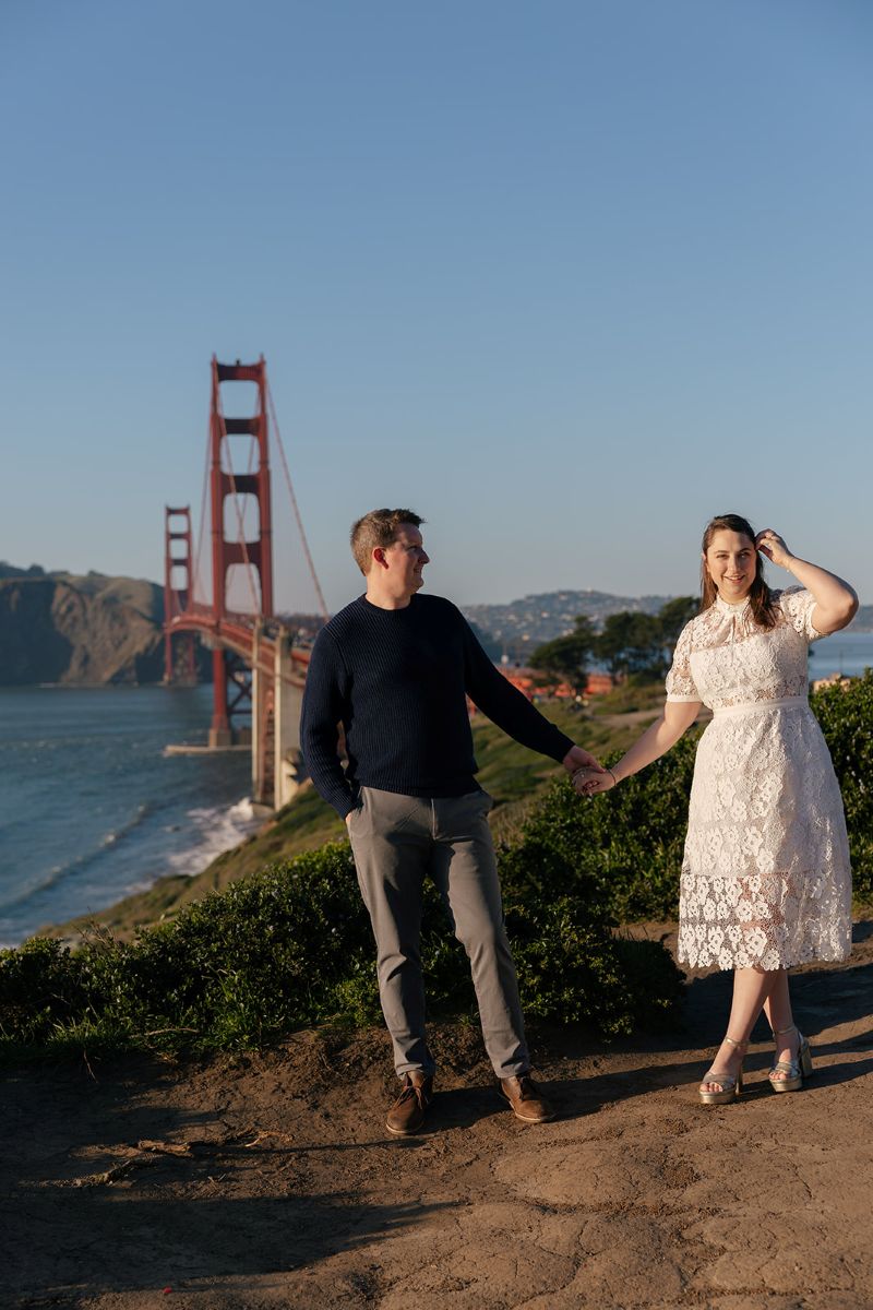 a couple taking engagement photos at battery godfrey in san francisco they are holding hands and the golden gate bridge is behind them