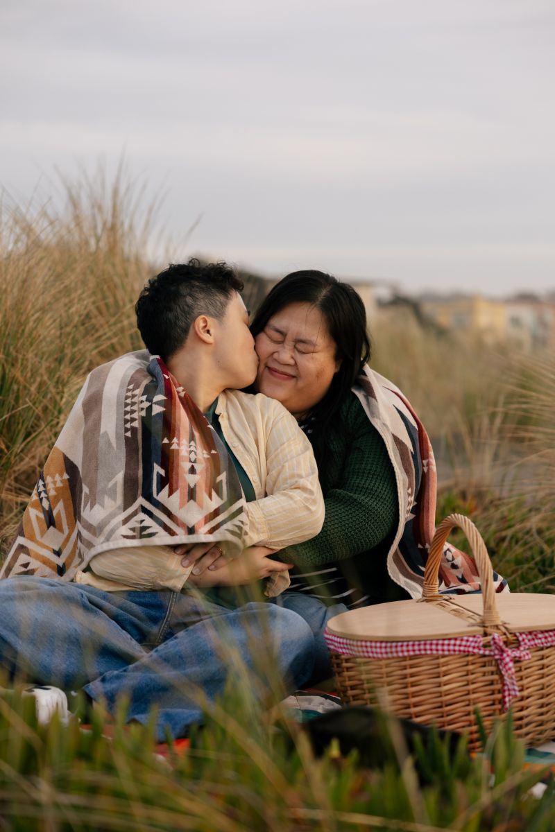 a couple having a picnic together next to the ocean one is kissing the other on the cheek