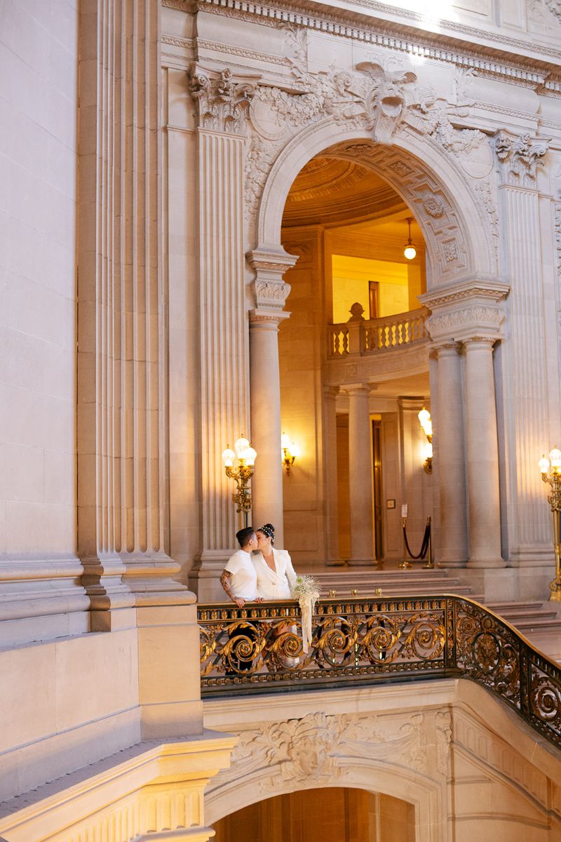 a couple taking engagement photos inside of san francisco city hall they are standing next to the banister and kissing one another