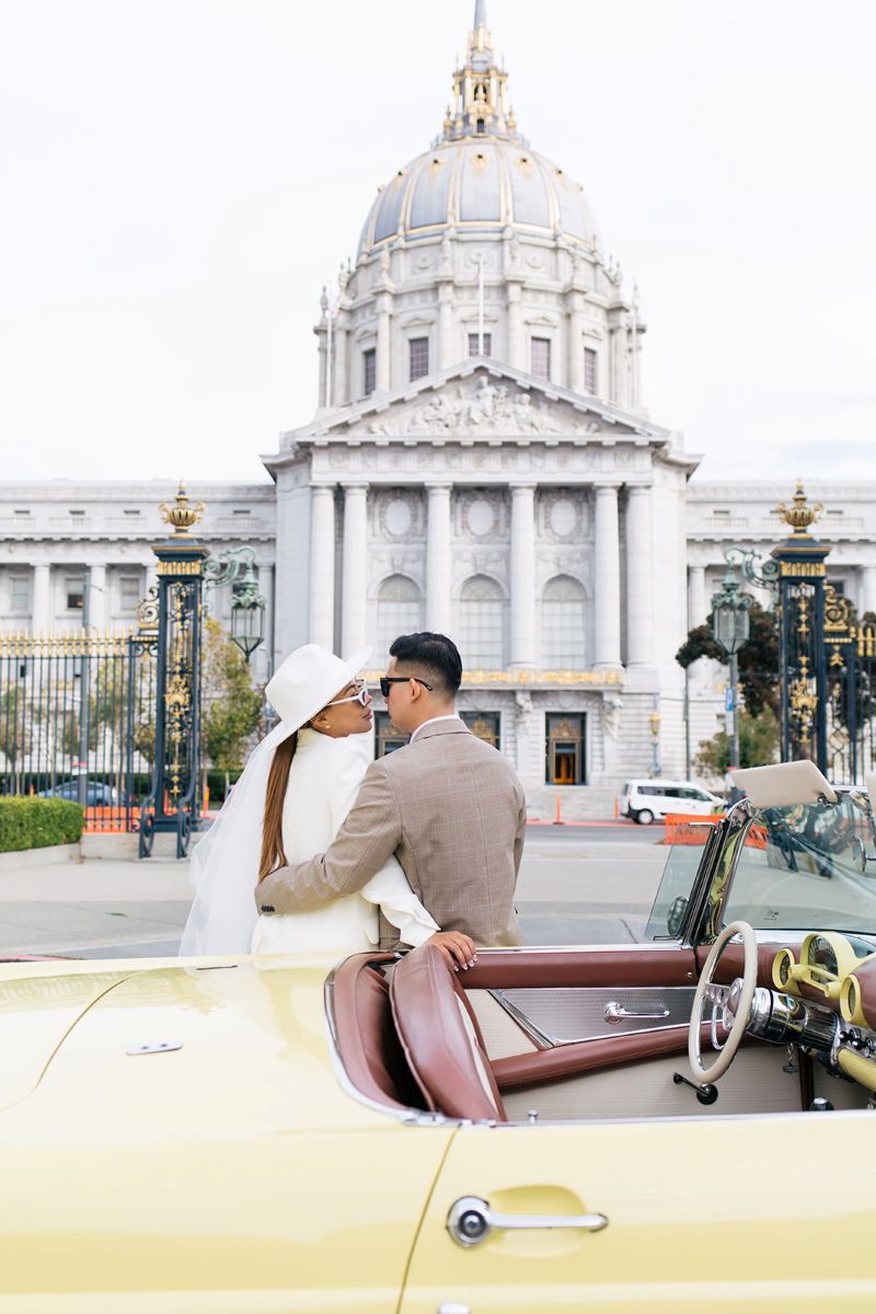 a couple having a vintage car engagement session in san francisco with a yellow car the woman is wearing a white suit, hat, and veil the man is wearing a tan suit
