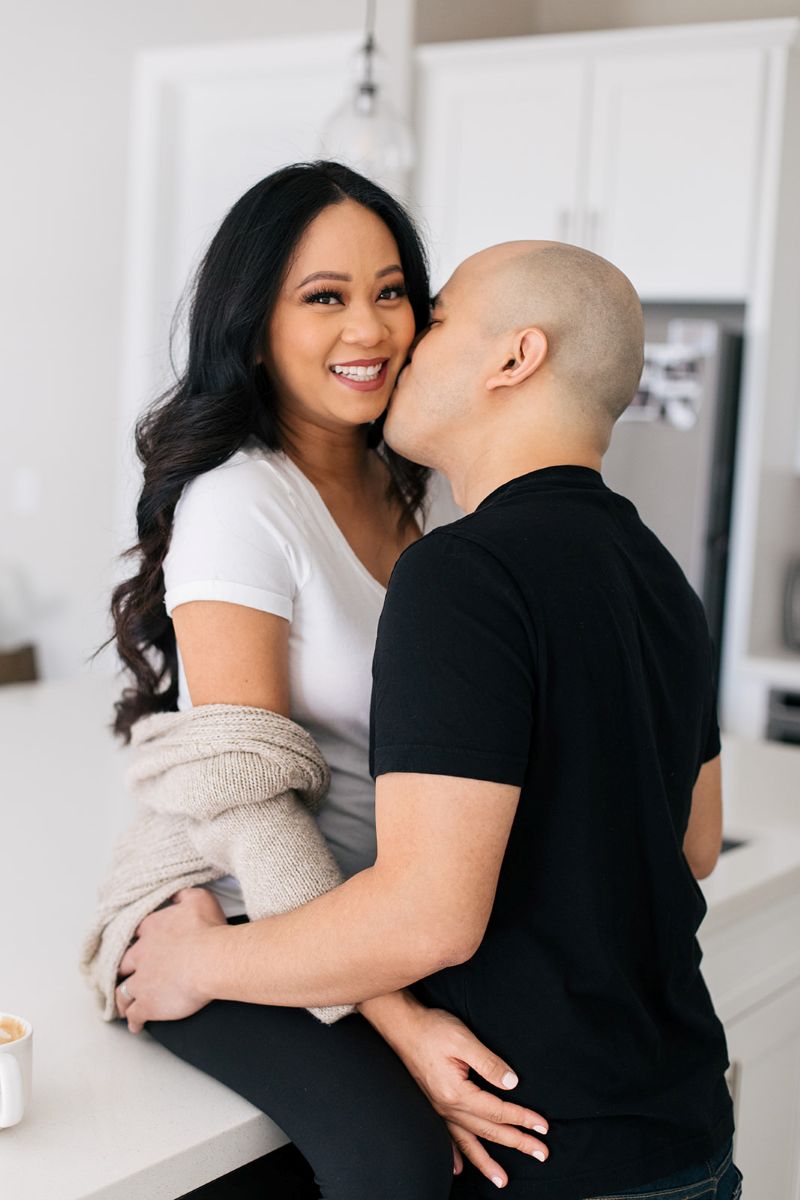 a couple having a lifestyle engagment session in their home the woman is sitting on the counter and her partner is kissing her on the cheek