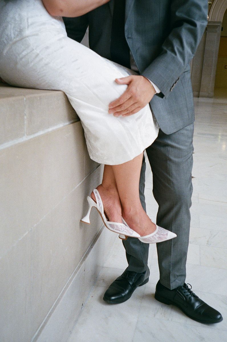 a woman sitting on a wall wearing a white wedding dress and heels and her partner is wearing a gray suit and touching her leg 