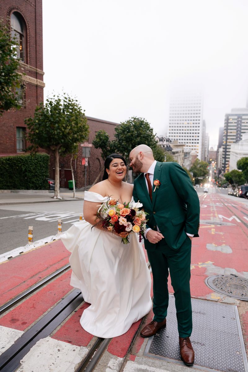 a couple laughing and crossing the tram tracks in san francisco the woman is wearing a white wedding dress and holding flowers the man is wearing a green suit 