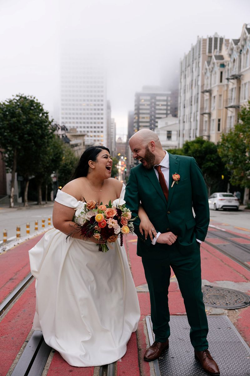 a couple laughing with each other with the city of san francisco behind them they are taking wedding photos 