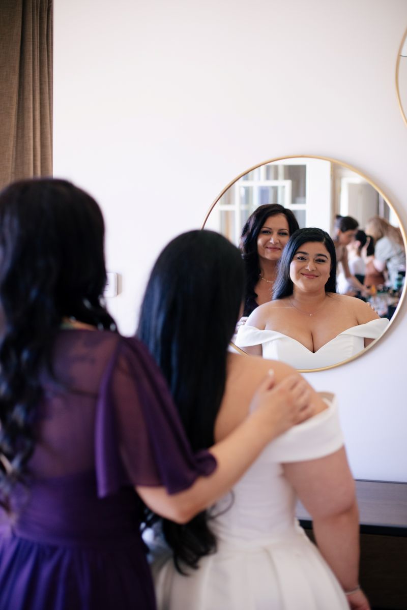 a woman wearing a white wedding dress smiling at a mirror with her mom touching her shoulder 