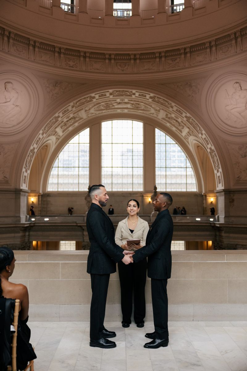 a couple having their wedding ceremony on the 4th floor of san francisco city hall they are holding hands and standing in front of their officiant