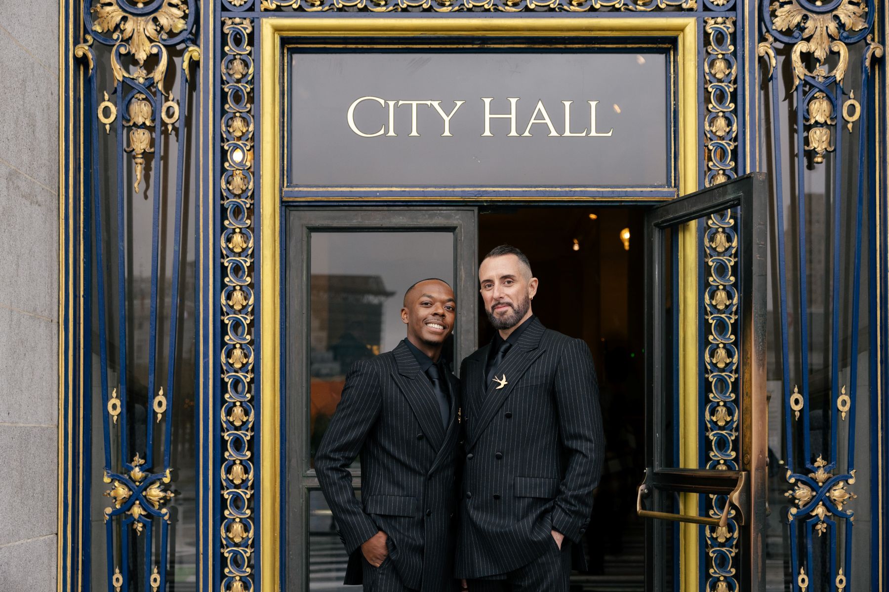 a couple in front of san francisco city hall on the day of their wedding standing directly in front of the entrance weraing matching black suits