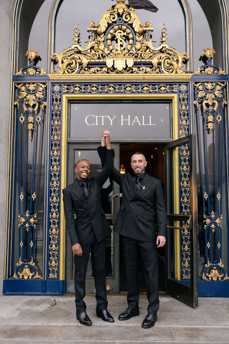 a couple holding their hands in the air in front of san francisco city hall they are both wearing matching black suits