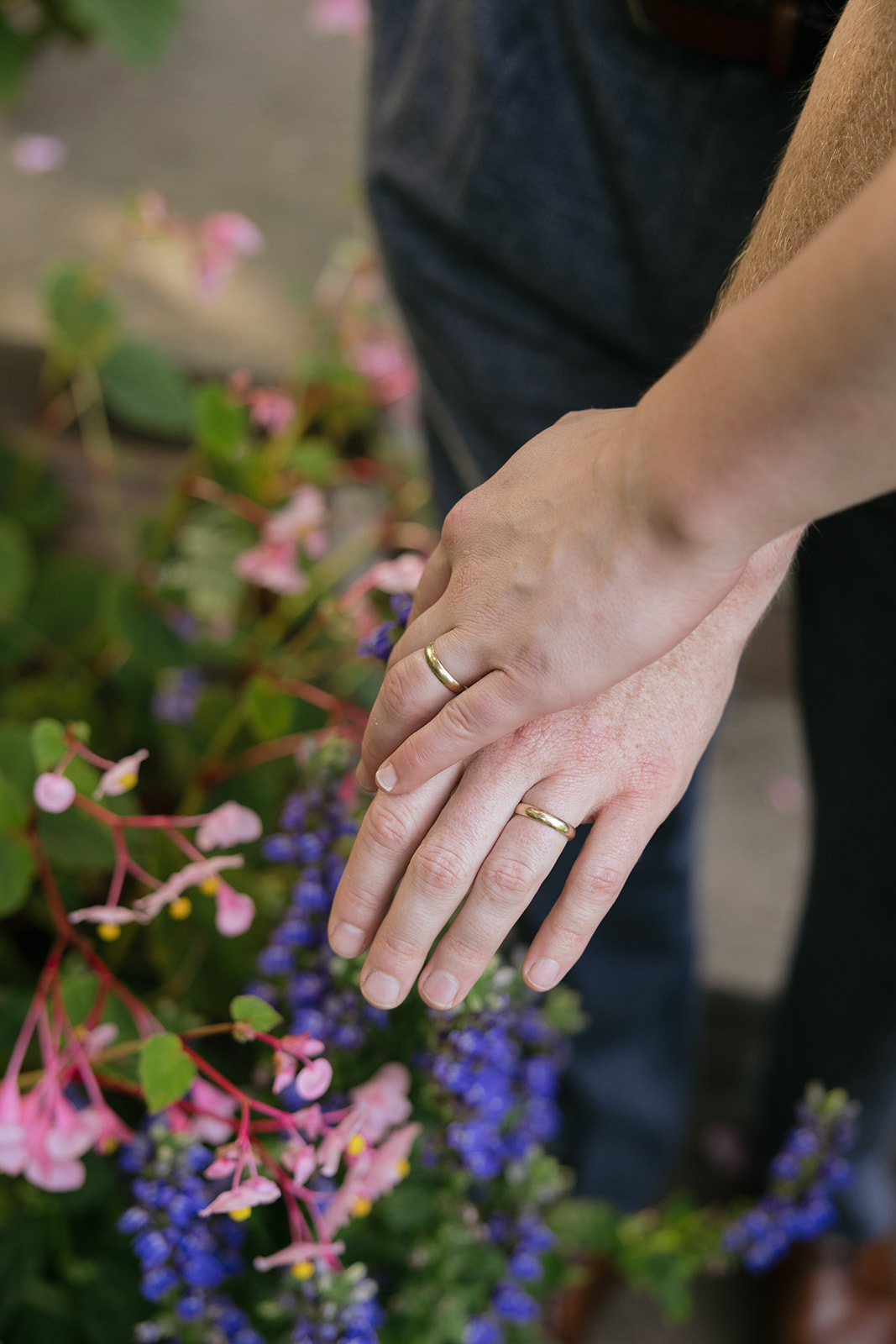 a close up photo of a couple's hands with engagement bands on top of each other with flowers in the background