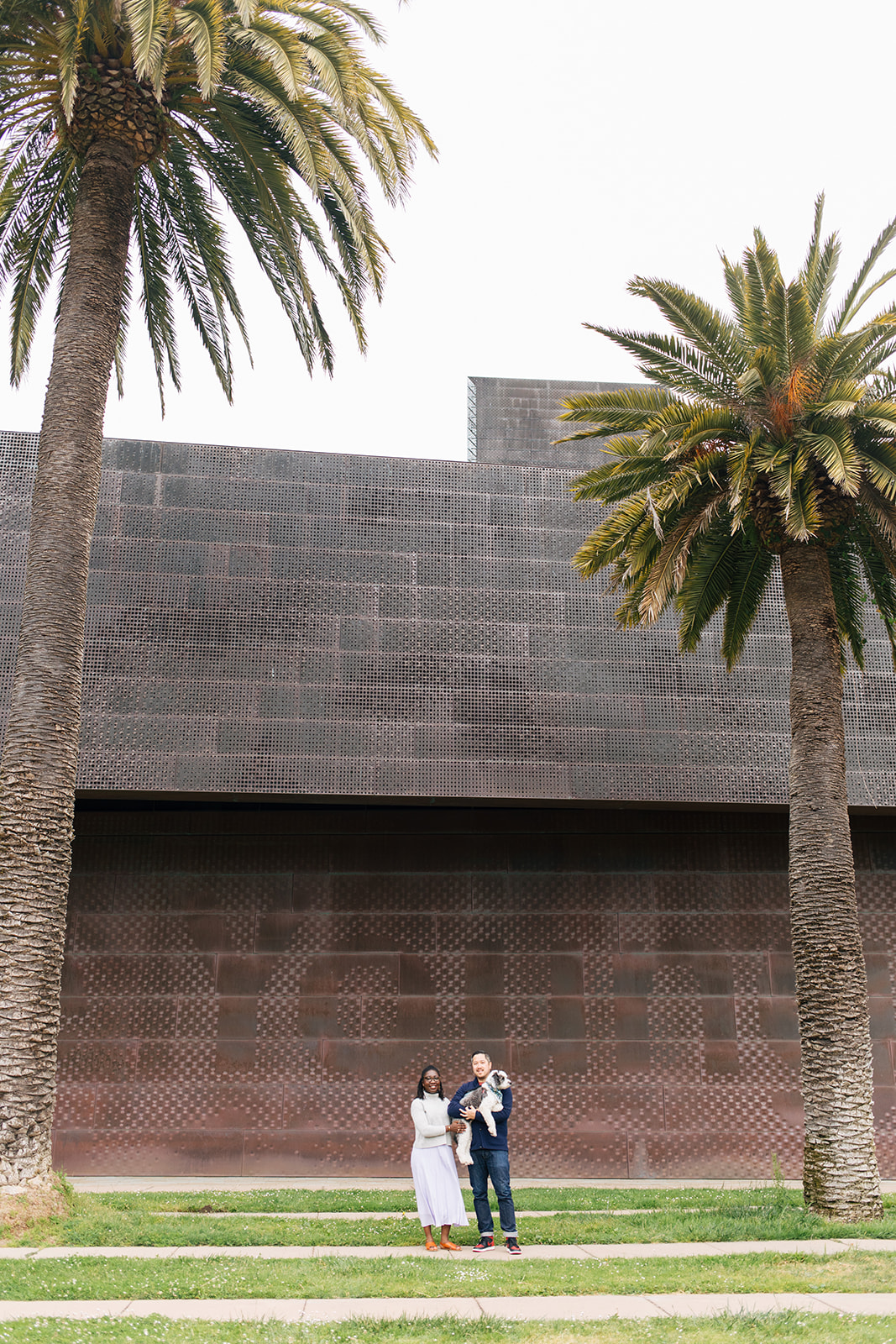 a couple and their dog standing in front of the de young museum in between two palm trees