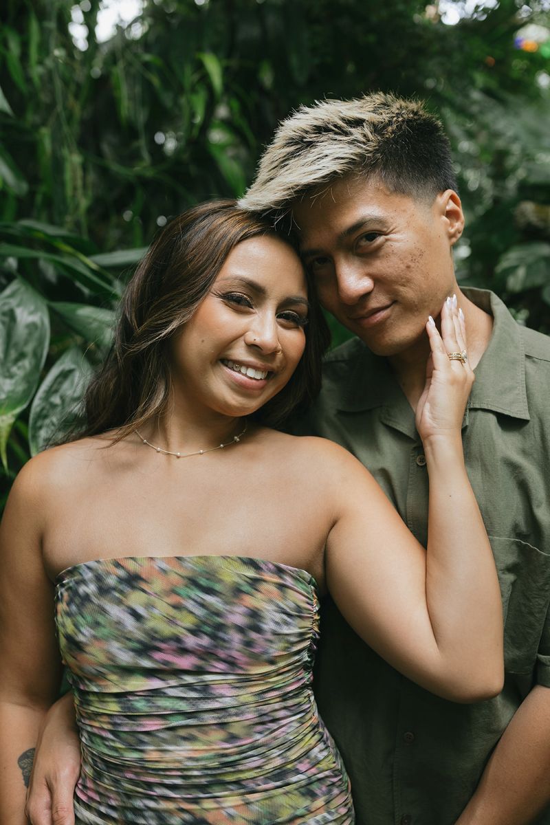 a couple taking engagement pictures in front of a variety of green plants inside of the Conservatory of Flowers in golden gate park the woman has her hand resting on her partner's cheek and they are both smiling 