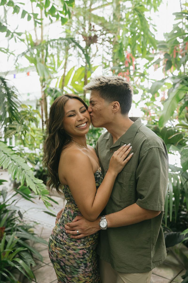 a couple taking engagement pictures in front of a variety of green plants inside of the Conservatory of Flowers in golden gate park the man is leaning towards his partner and kissing her cheek and hugging her