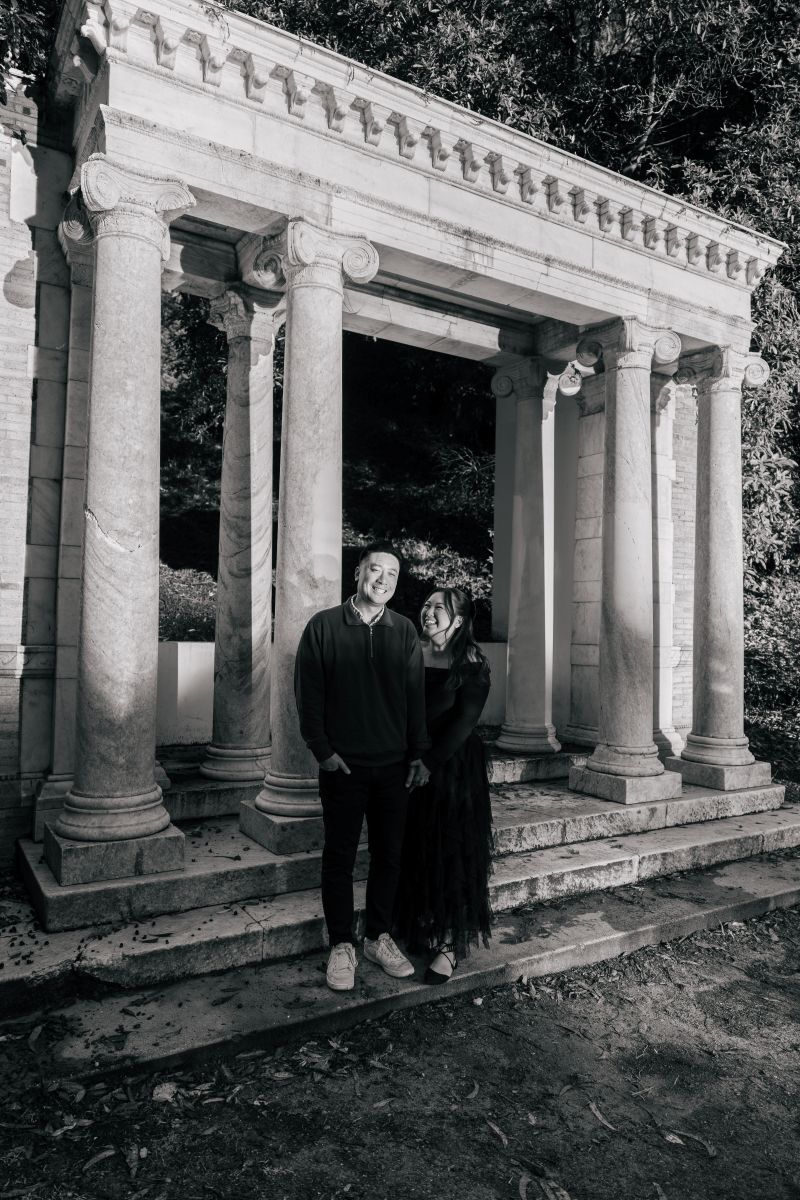 a couple taking engagement pictures at portals of the past inside of golden gate park they are both holding hands and standing in front of marble columns 