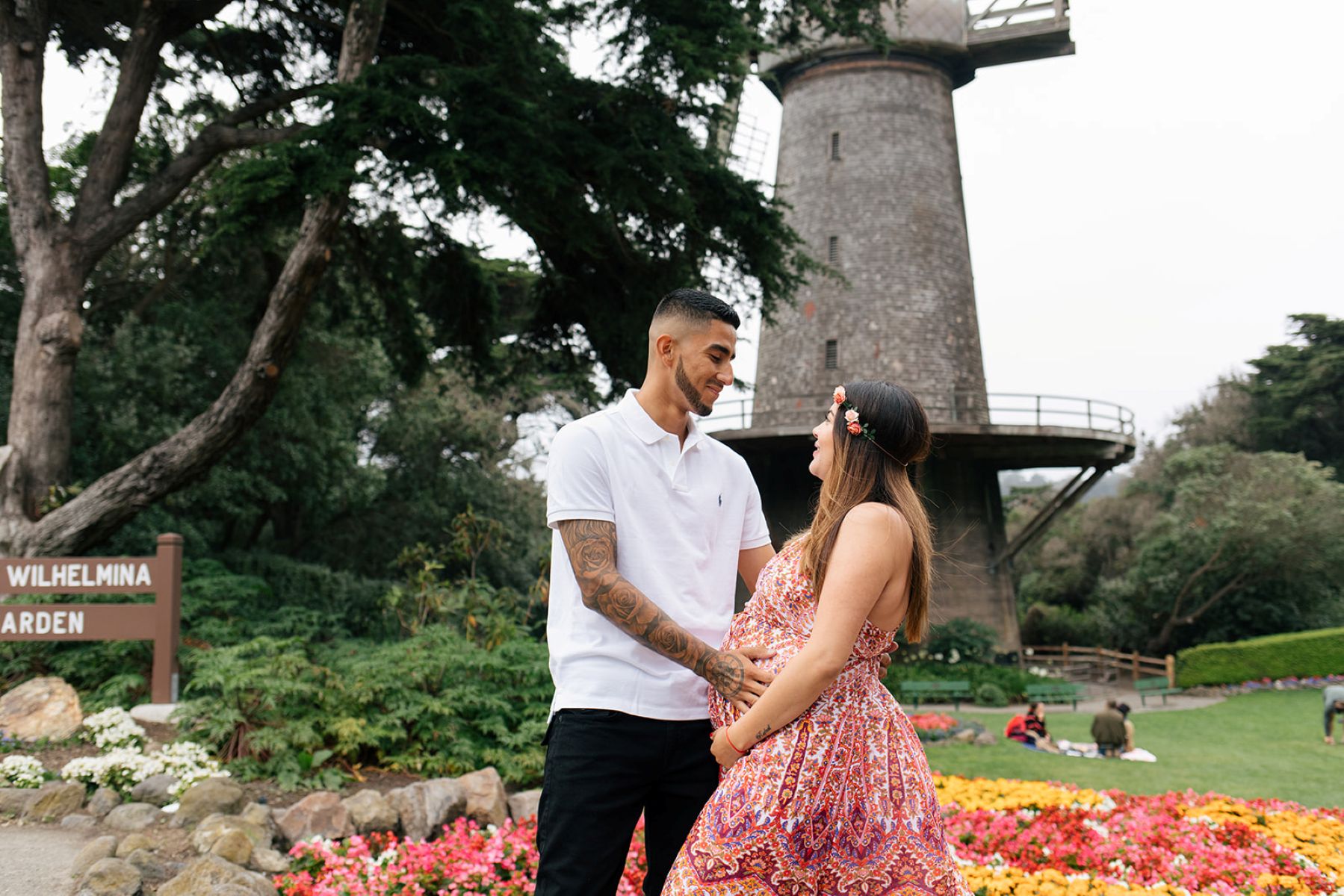 a couple taking maternity pictures at Queen Wilhelmina Tulip Garden inside of golden gate park the man is touching his partner on her stomach and she is smiling at him behind them are a variety of flowers 