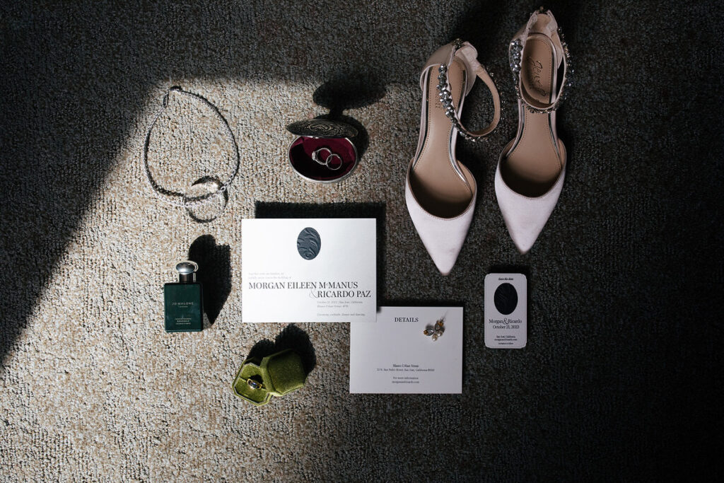 A wedding flat lay photo showing invitations, perfume, jewelry, rings, and white heels 