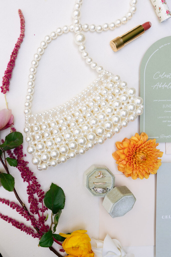 A pearl handbag, rings in a ring box, flowers, wedding invitations, and lipstick laid out together 