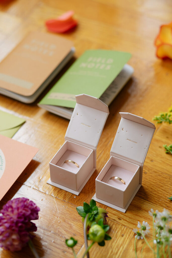 Two matching ring boxes with wedding bands in them on a table with small notebooks and florals 