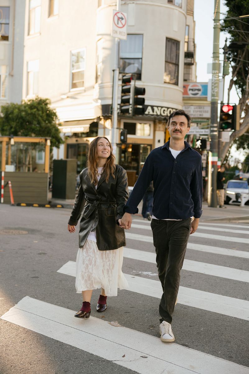 a couple walking across a sidewalk in north beach holding hands and smiling at each other