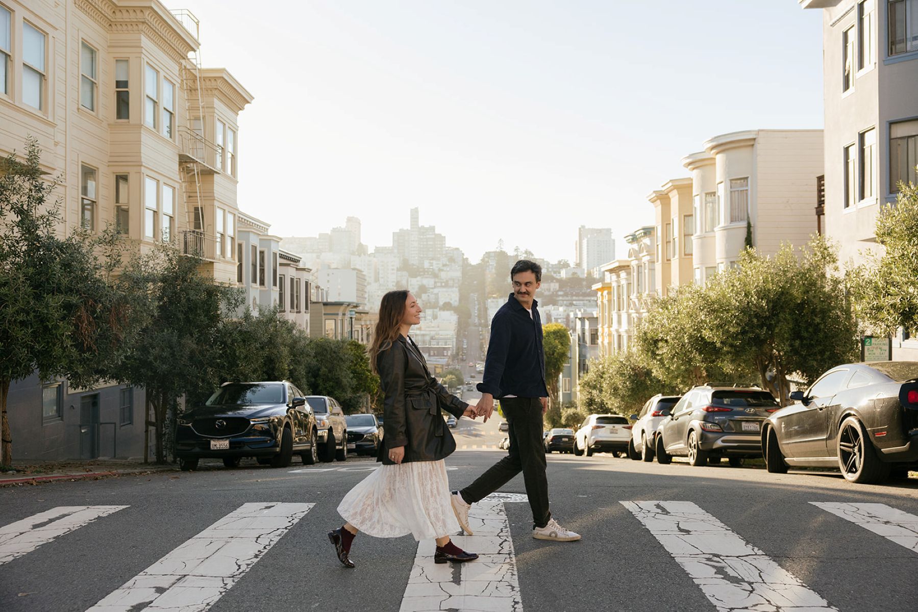 a couple walking hand in hand during their san francisco engagement session across a sidewalk with the san francisco hills behind them