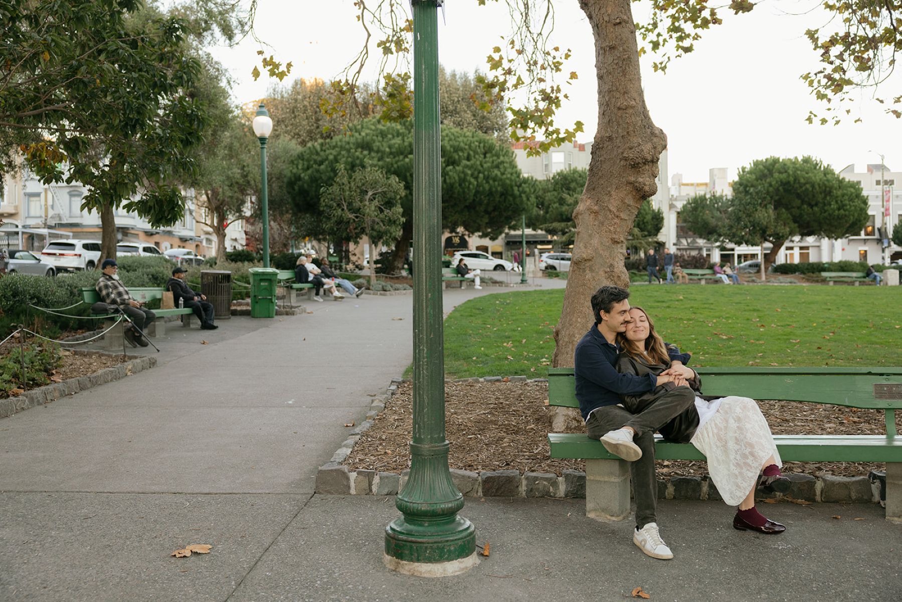 a couple sitting on a green park bench next to a park in north beach the man has his arms wrapped around his partner and she is leaning back into him