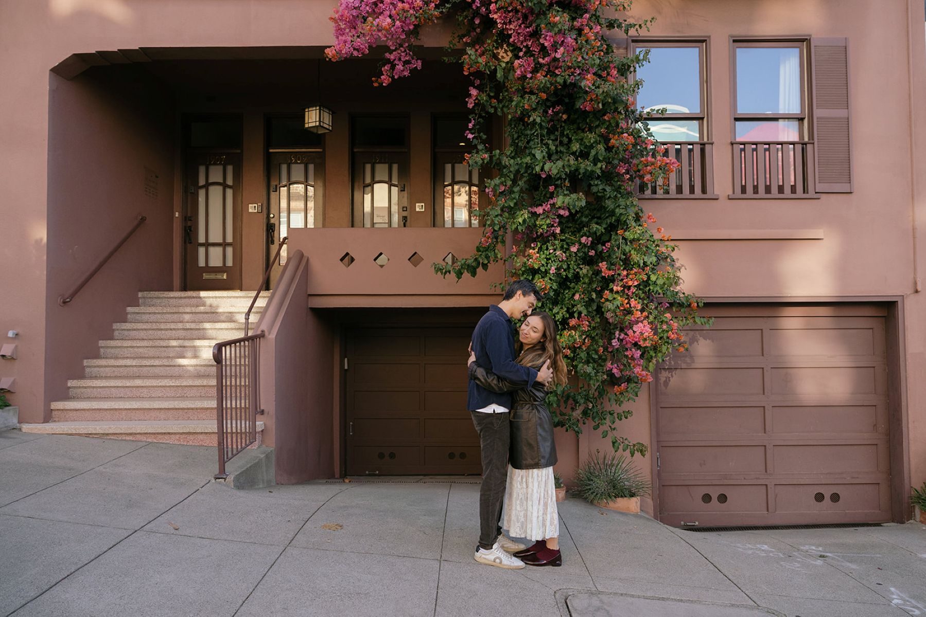 a man hugging his partner in front of a pink house and a bougainvillea vine