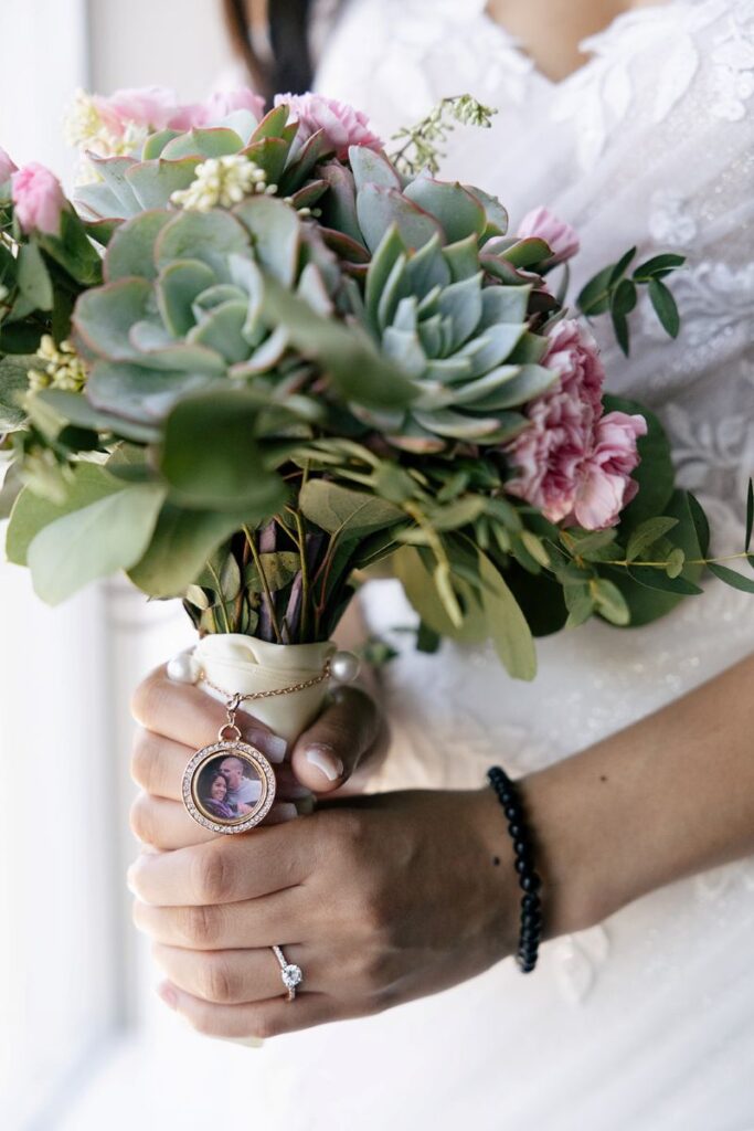 a bride holding a bouquet next to a window the bouquet has a small charm on it with a picture inside 