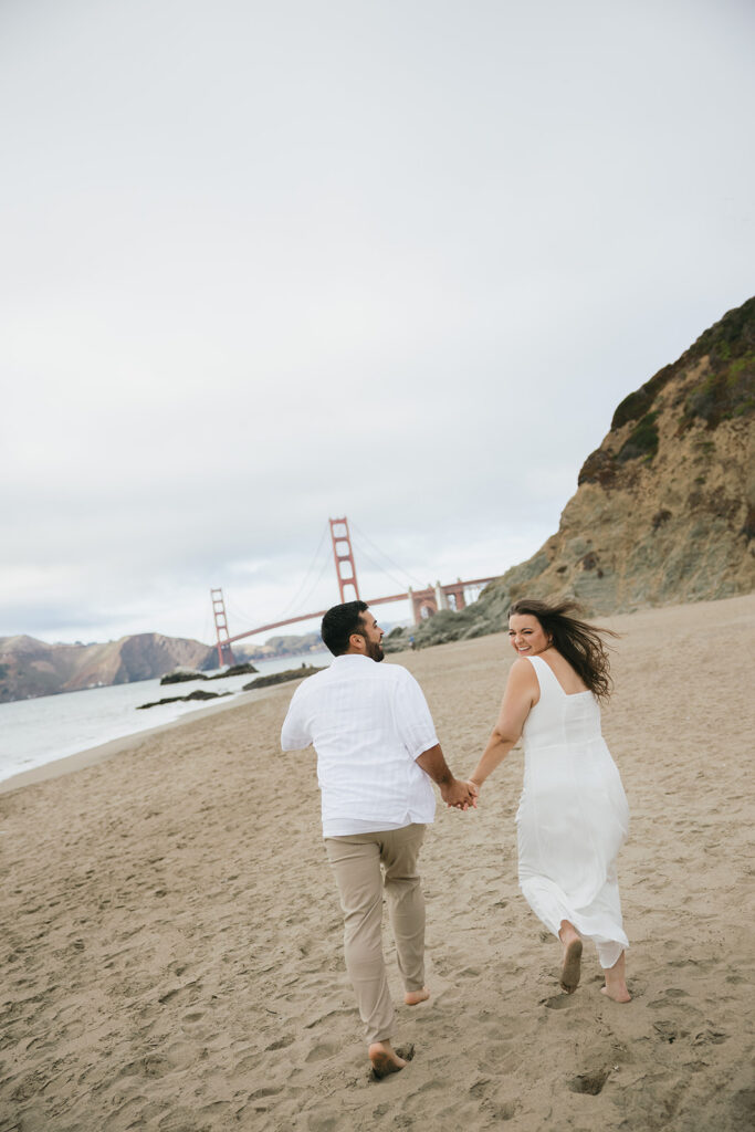 a couple holding hands and running on the beach towards the golden gate bridge 
